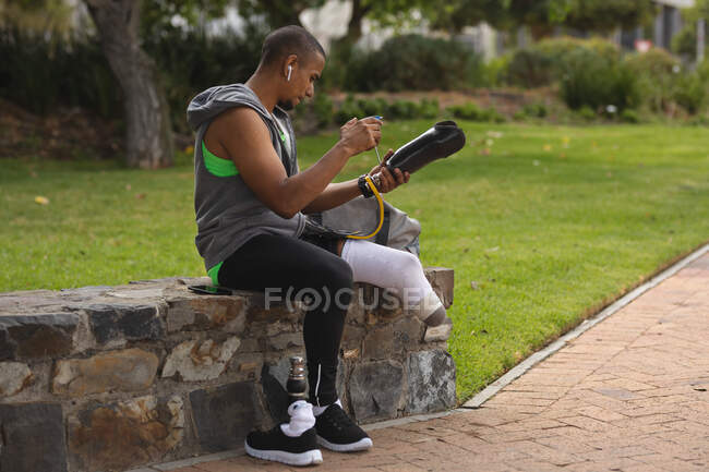 Homme handicapé de race mixte avec une jambe prothétique, travaillant dans un parc urbain, assis sur un mur par un chemin faisant des ajustements à sa jambe prothétique. Fitness handicap mode de vie sain. — Photo de stock