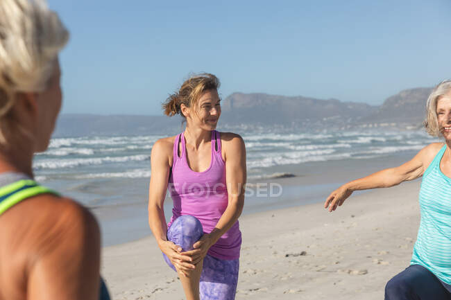 Groupe d'amies caucasiennes profitant de l'exercice sur une plage par une journée ensoleillée, pratiquant le yoga et s'étirant en position de yoga. — Photo de stock