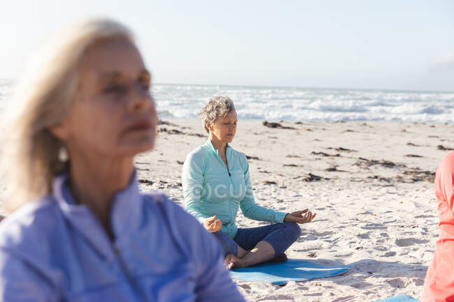 Group of Caucasian female friends enjoying exercising on a beach on a sunny day, practicing yoga, meditating in lotus position, with sea in the background. — Stock Photo