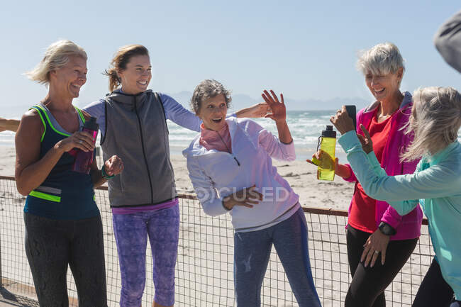 Grupo de amigas caucasianas que gostam de se exercitar em uma praia em um dia ensolarado, fazer uma pausa, de pé em um passeio marítimo e tirar fotos com um smartphone. — Fotografia de Stock