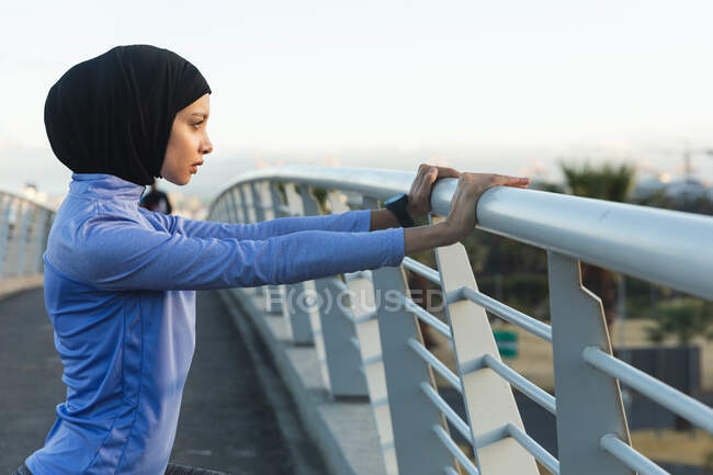 Fit gemischte Rennläuferin in Hijab und Sportbekleidung, die an einem sonnigen Tag im Freien in der Stadt trainiert und sich auf einer Fußgängerbrücke dehnt. Bewegung im städtischen Lebensstil. — Stockfoto