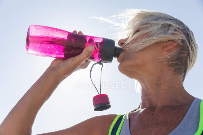 Ältere kaukasische Frau genießt an einem sonnigen Tag das Training am Strand, ruht sich aus, nachdem sie am Strand gelaufen ist und Wasser getrunken hat. — Stockfoto