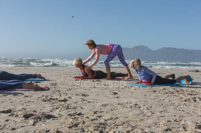 Grupo de amigas caucásicas disfrutando haciendo ejercicio en una playa en un día soleado, practicando yoga y estirándose en posición de yoga. - foto de stock