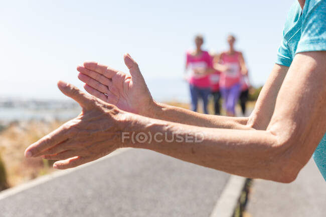 Manos caucásicas grupo motivador de amigas caucásicas disfrutando de hacer ejercicio en un día soleado, correr, usar números y ropa deportiva rosa. - foto de stock