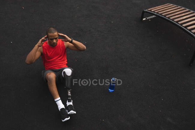 Hombre de raza mixta discapacitado con una pierna protésica, haciendo ejercicio en un parque, escuchando música en los auriculares, haciendo abdominales. Fitness discapacidad estilo de vida saludable. - foto de stock