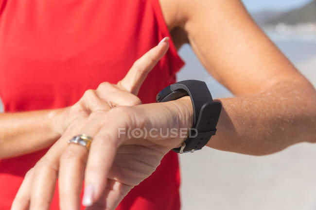 Primer plano de la mujer disfrutando de hacer ejercicio en una playa en un día soleado, de pie y usando su smartwatch con el mar en el fondo. - foto de stock