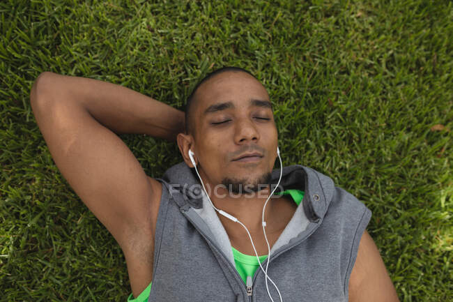Hombre de raza mixta usando ropa deportiva, haciendo ejercicio en un parque, tomando un descanso acostado en la hierba con los ojos cerrados usando auriculares y escuchando música. Fitness estilo de vida saludable. - foto de stock