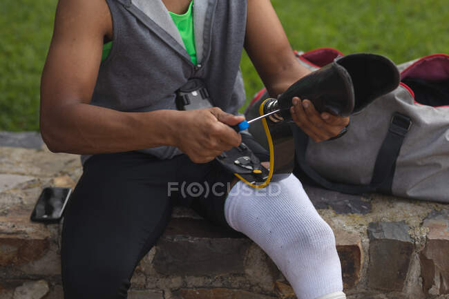 Partie médiane d'un homme handicapé avec une jambe prothétique travaillant dans un parc urbain, assis sur un mur et ajustant une lame de course. Fitness handicap mode de vie sain. — Photo de stock
