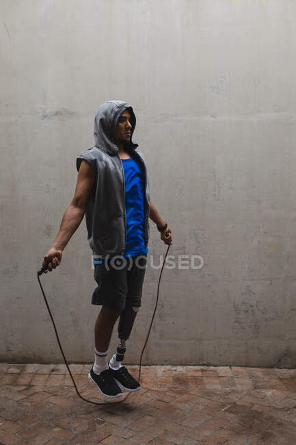 Uomo disabile di razza mista con una gamba protesica, che si allena in un parco urbano, indossa un top con cappuccio che salta con corda per saltare. Fitness disabilità stile di vita sano. — Foto stock