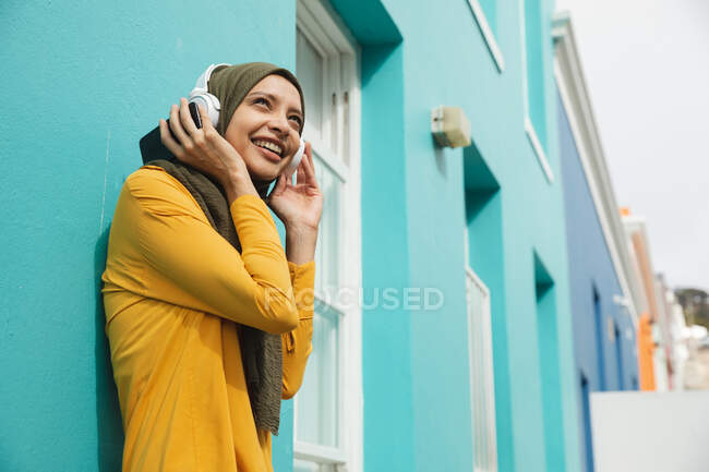 Femme de race mixte portant hijab et pull jaune sur et sur la route dans la ville, souriant avec des écouteurs sans fil sur le support contre le mur bleu. Commuter style de vie moderne. — Photo de stock