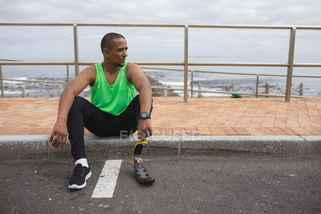 Disabled mixed race man with a prosthetic leg and running blade working out by the coast, sitting on the pavement by a road and taking a break. Fitness disability healthy lifestyle. — Stock Photo