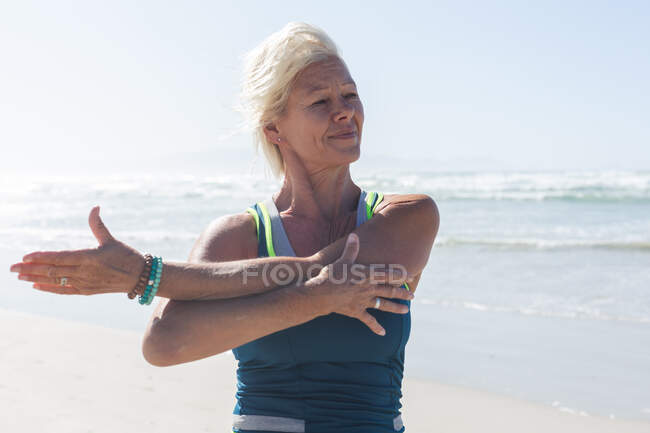 Ältere kaukasische Frau mit blonden Haaren genießt an einem sonnigen Tag das Training am Strand, praktiziert Yoga und dehnt sich mit dem Meer im Hintergrund. — Stockfoto