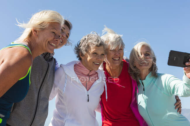 Group of Caucasian female friends enjoying exercising on a beach on a sunny day, taking photo with a smartphone, smiling and embracing. — Stock Photo