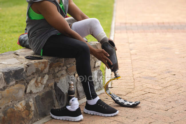 Низкая часть инвалида с протезной ногой работает в городском парке, сидит на стене и устанавливает бегущее лезвие. Здоровый образ жизни. — стоковое фото