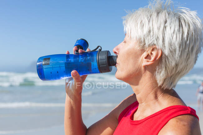 Donna caucasica anziana che si diverte ad allenarsi su una spiaggia in una giornata di sole, riposandosi dopo aver corso sulla riva del mare e bevendo acqua da una bottiglia. — Foto stock