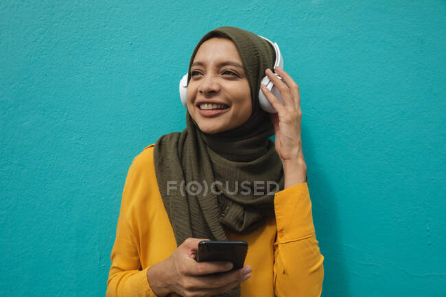 Donna di razza mista che indossa hijab e maglione giallo in giro per la città, sorridente tenendo smartphone con cuffie wireless. Stile di vita moderno pendolare. — Foto stock