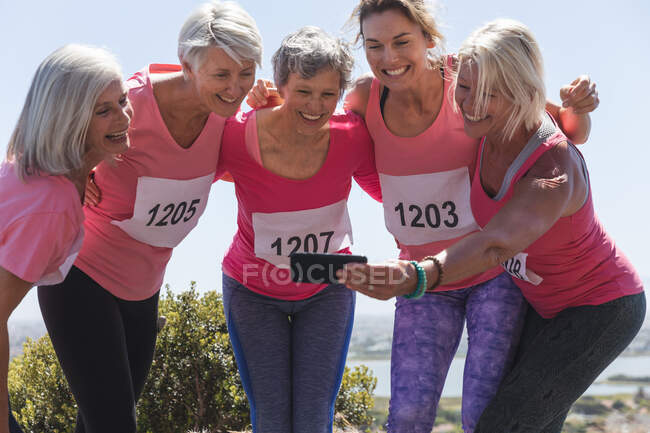 Grupo de amigas caucásicas disfrutando haciendo ejercicio en un día soleado, celebrando después de correr, llevando números y sonriendo, tomando una foto con smartphone. - foto de stock