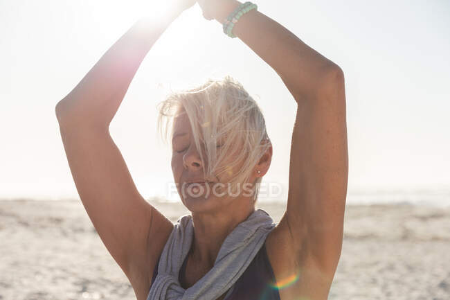 Femme caucasienne âgée qui aime faire de l'exercice sur une plage par une journée ensoleillée, pratiquer le yoga, debout en position d'arbre les yeux fermés. — Photo de stock