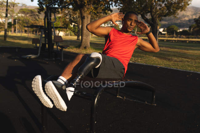 Uomo disabile di razza mista con una gamba protesica che indossa abbigliamento sportivo, si allena in un parco, ascolta musica sugli auricolari, fa crunches. Fitness disabilità stile di vita sano. — Foto stock