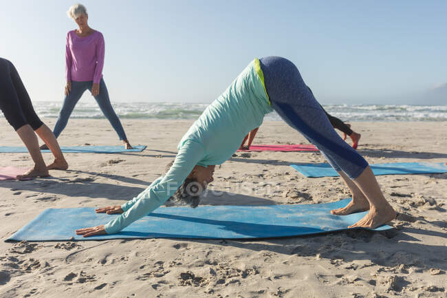 Groupe d'amies caucasiennes profitant de l'exercice sur une plage par une journée ensoleillée, pratiquant le yoga et se tenant debout en position de chien. — Photo de stock