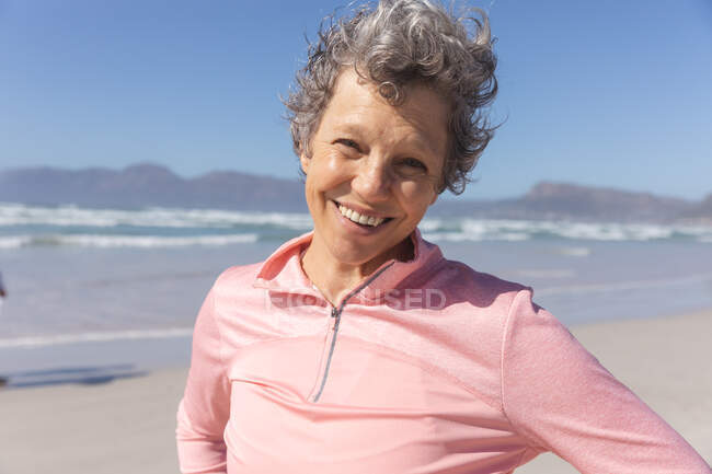 Porträt einer älteren kaukasischen Frau, die es genießt, an einem sonnigen Tag am Strand Sport zu treiben, lächelt, steht und in die Kamera schaut, im Hintergrund das Meer. — Stockfoto