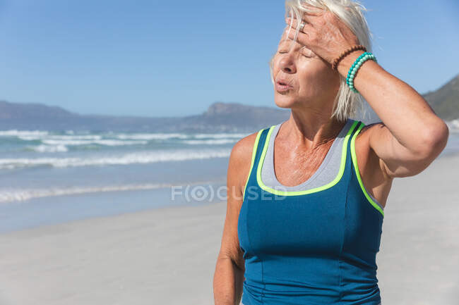 Старшая белая женщина, занимающаяся спортом на пляже в солнечный день, отдыхающая после пробежки по берегу моря и прикосновения ко лбу. — стоковое фото