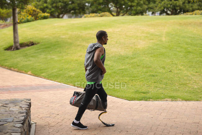 Homme handicapé de race mixte avec une jambe prothétique travaillant dans un parc urbain, portant une lame de course, portant un sac et marchant sur un chemin. Fitness handicap mode de vie sain. — Photo de stock