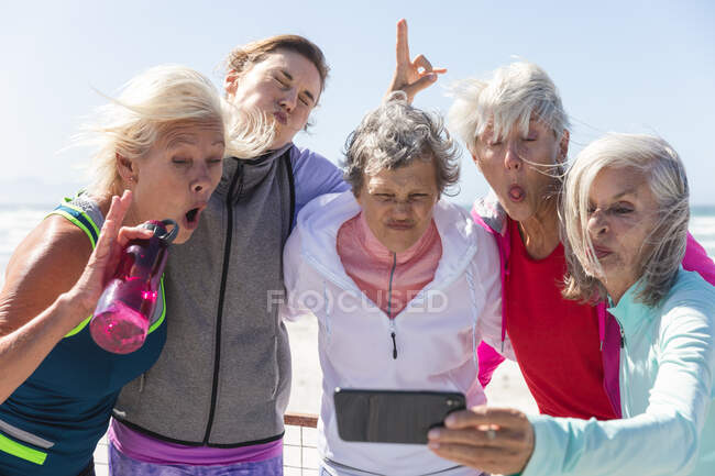 Группа кавказских подруг, занимающихся спортом на пляже в солнечный день, улыбающихся, фотографирующихся со смартфоном. — стоковое фото