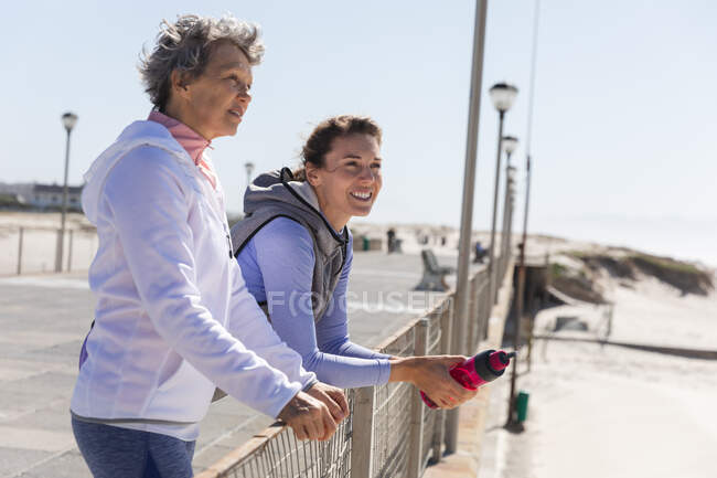 Две кавказские подружки наслаждаются тренировками на пляже в солнечный день, улыбаясь, стоя на набережной с морем на заднем плане. — стоковое фото