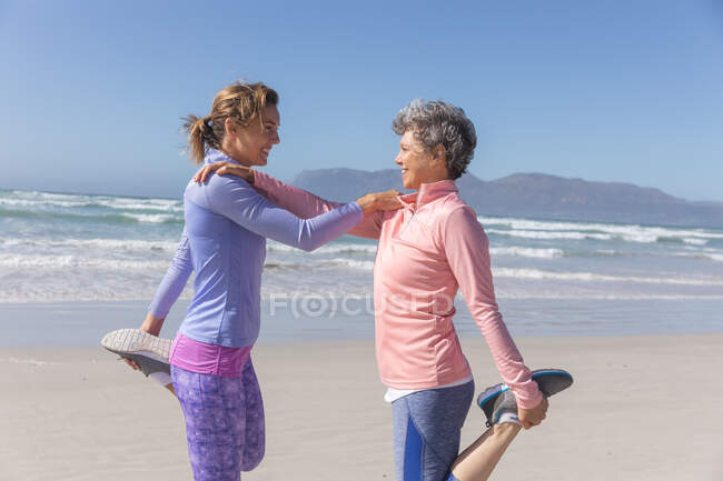 Dos amigas caucásicas disfrutando haciendo ejercicio en una playa en un día soleado, practicando yoga y estirándose con el mar al fondo. - foto de stock