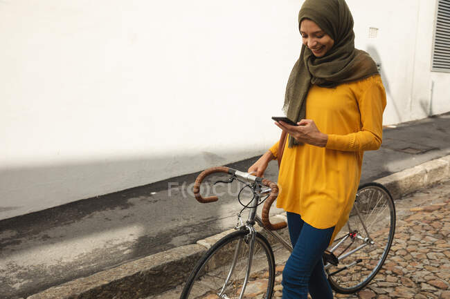 Donna di razza mista che indossa hijab e maglione giallo in giro per la città, utilizzando il suo smartphone sorridente a piedi con la bicicletta. Stile di vita moderno pendolare. — Foto stock