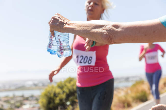 Caucasian hand giving senior Caucasian woman a bottle of water during running race, enjoying exercising on a sunny day, wearing numbers and pink sportswear. — Stock Photo