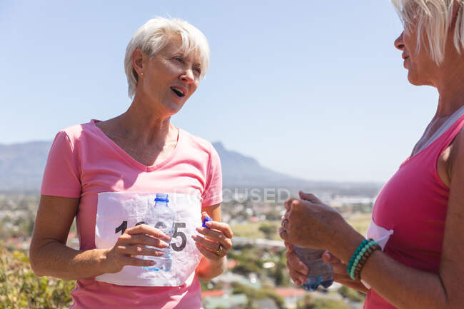 Dos amigas caucásicas mayores disfrutando haciendo ejercicio en un día soleado, tomando un descanso después de correr, usando números y ropa deportiva rosa, bebiendo agua de una botella. - foto de stock