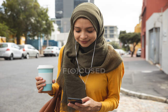 Gemischte Frau in Hijab und gelbem Pullover unterwegs in der Stadt, Kaffee zum Mitnehmen per Smartphone mit Kopfhörer in der Hand. Pendler moderner Lebensstil. — Stockfoto