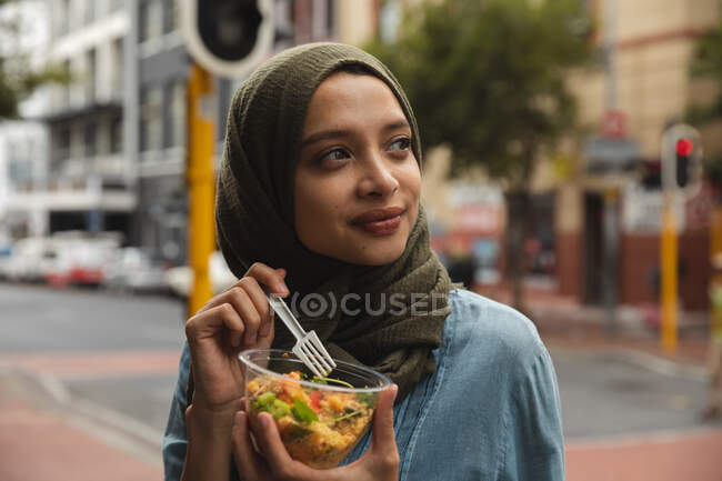 Mujer de raza mixta con hijab fuera y sobre la marcha en la ciudad, de pie en la calle comiendo comida para llevar sosteniendo tazón y tenedor, sonriendo. Commuter estilo de vida moderno. - foto de stock