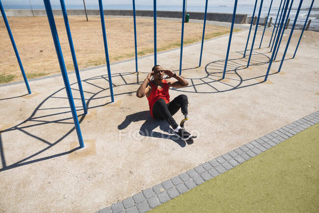 Hombre de raza mixta discapacitado con una pierna protésica y una cuchilla corriendo ejercitándose en un gimnasio al aire libre junto a la costa, haciendo sentadas al sol junto al equipo del gimnasio. Fitness discapacidad estilo de vida saludable. - foto de stock