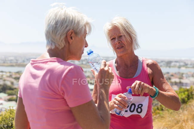 Zwei ältere kaukasische Freundinnen genießen das Training an einem sonnigen Tag, machen nach dem Lauf eine Pause, tragen Nummern und rosa Sportbekleidung, trinken Wasser aus einer Flasche. — Stockfoto