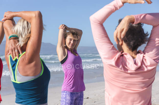 Grupo de amigas caucasianas que gostam de se exercitar em uma praia em um dia ensolarado, praticando ioga e alongamento com o mar no fundo. — Fotografia de Stock