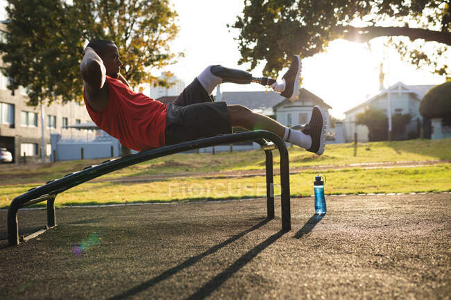 Disabled mixed race man with a prosthetic leg wearing sportswear, working out in a park, doing crunches on bench. Fitness disability healthy lifestyle. — Stock Photo