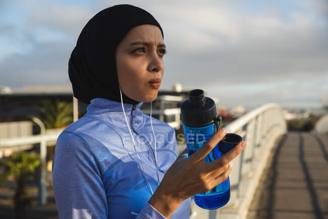 Fit mixte femme de race portant hijab et vêtements de sport exercice à l'extérieur dans la ville par une journée ensoleillée, tenant bouteille d'eau prendre une pause en portant des écouteurs sur une passerelle. Exercice mode de vie urbain. — Photo de stock