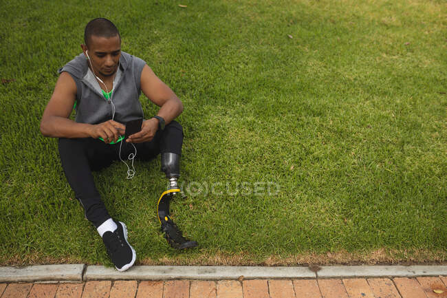 Неповносправний гоночний чоловік з протезом ноги і лезо, що працює в парку, робить перерву, сидить на траві за допомогою смартфона і носить навушники. Здоровий спосіб життя - інвалідність. — стокове фото