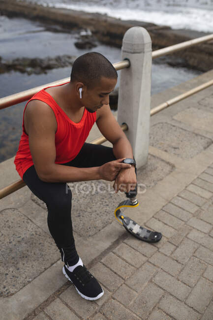 Behinderter Mixed Racer mit Beinprothese und Laufmesser, der mit drahtlosen Kopfhörern an der Küste trainiert, auf einem Zaun sitzt und die Smartwatch kontrolliert. Fitness Behinderung gesunder Lebensstil. — Stockfoto