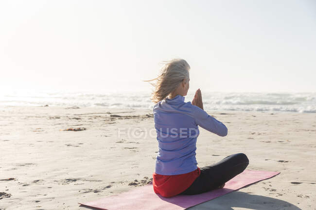 Mujer caucásica con cabello rubio disfrutando haciendo ejercicio en una playa en un día soleado, practicando yoga y sentada en posición de yoga. - foto de stock