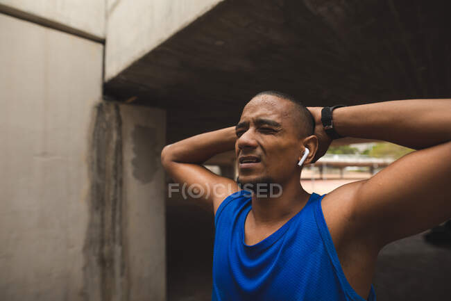 Homme de race mixte travaillant dans un parc urbain, portant des vêtements de sport, smartwatch et écouteurs sans fil, clignotant avec douleur et étirement. Fitness force mode de vie sain. — Photo de stock