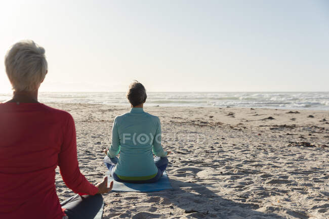 Grupo de amigas caucásicas disfrutando haciendo ejercicio en una playa en un día soleado, practicando yoga, meditando en posición de loto, frente al mar. - foto de stock