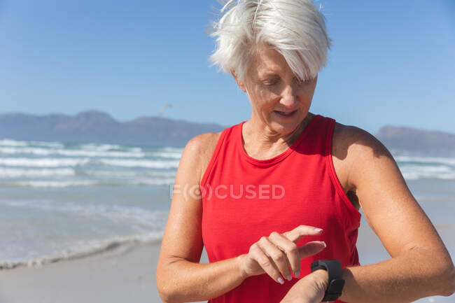 Ältere kaukasische Frau genießt an einem sonnigen Tag das Training am Strand, steht und benutzt ihre Smartwatch mit dem Meer im Hintergrund. — Stockfoto