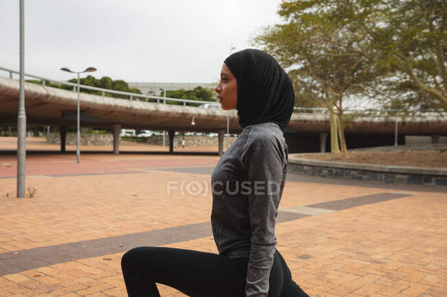 Fit mulher de raça mista vestindo hijab e sportswear exercitando ao ar livre na cidade, aquecendo-se alongamento no parque urbano. Exercício urbano. — Fotografia de Stock