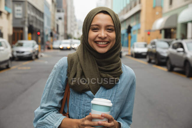 Retrato de una mujer de raza mixta con hijab fuera y sobre la marcha en la ciudad, de pie en la calle sosteniendo café para llevar, sonriendo a la cámara. Commuter estilo de vida moderno. - foto de stock