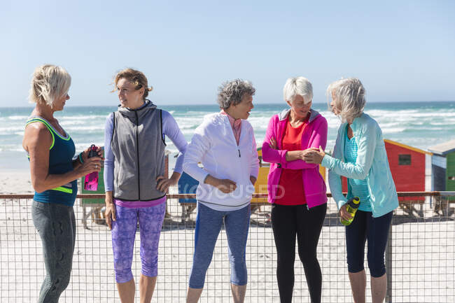 Grupo de uma amiga caucasiana que gosta de se exercitar em uma praia em um dia ensolarado, sorrindo, de pé em um passeio com pequenas casas coloridas no fundo. — Fotografia de Stock