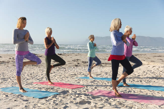Grupo de amigas caucásicas disfrutando haciendo ejercicio en una playa en un día soleado, practicando yoga, de pie en posición de árbol con el mar al fondo. - foto de stock